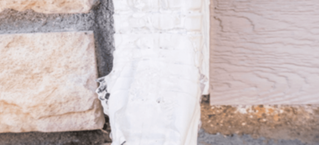 White sealant fills a gap between textured stone and wood siding on an exterior wall.