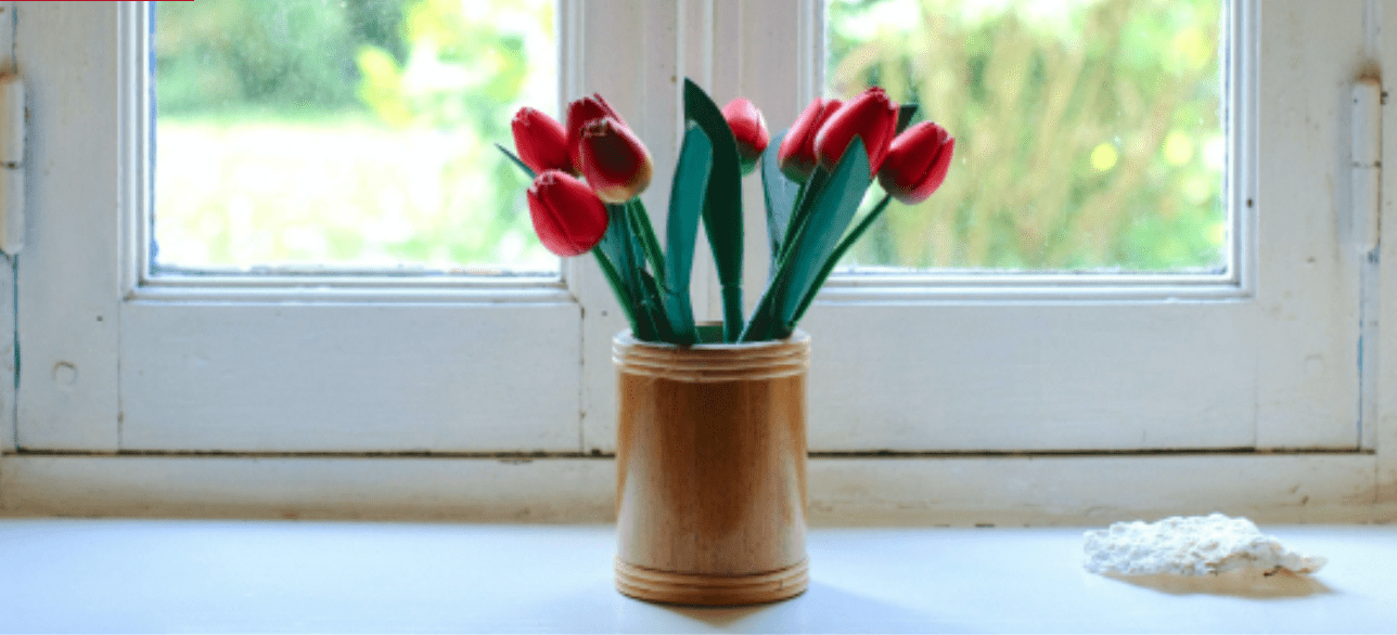 Red tulips in a wooden vase sit on a white windowsill overlooking a blurred green garden. TITLE