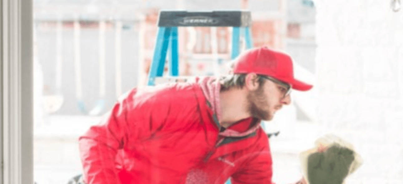A man in a red jacket and cap cleans a window with a blue ladder visible in the background