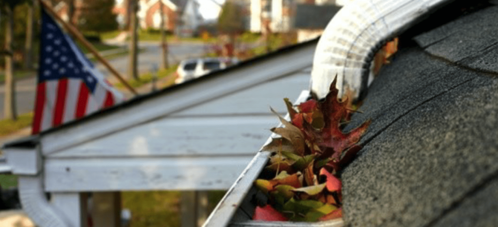 A white house gutter is overflowing with colorful autumn leaves, needing to be cleaned.