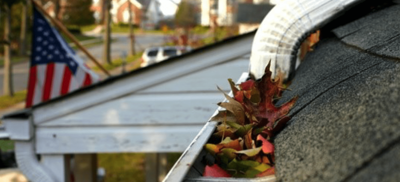 A white house gutter is overflowing with colorful autumn leaves, needing to be cleaned.