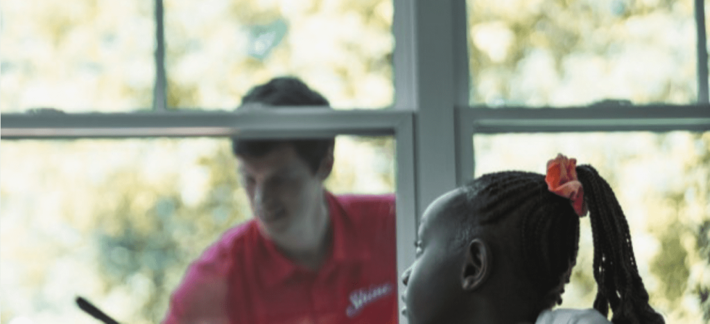 A young girl with braided hair watches a man in a red service shirt through a sunlit
