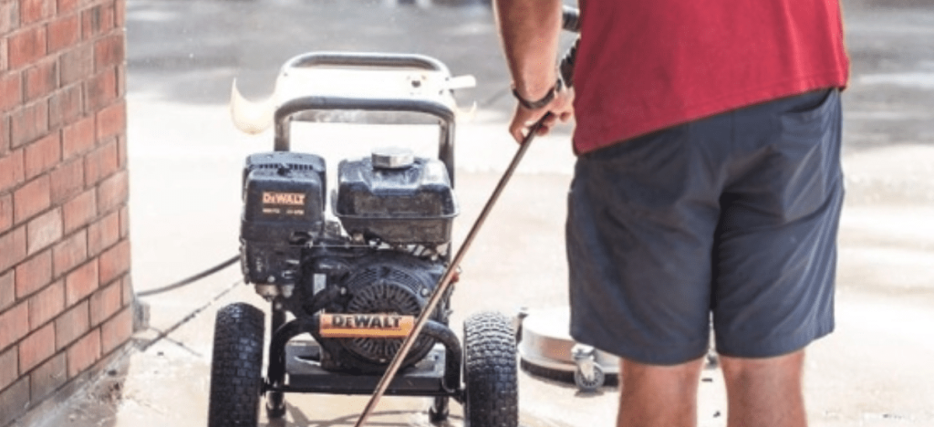 A person operates a DeWalt pressure washer to clean a wet surface next to a brick wall.