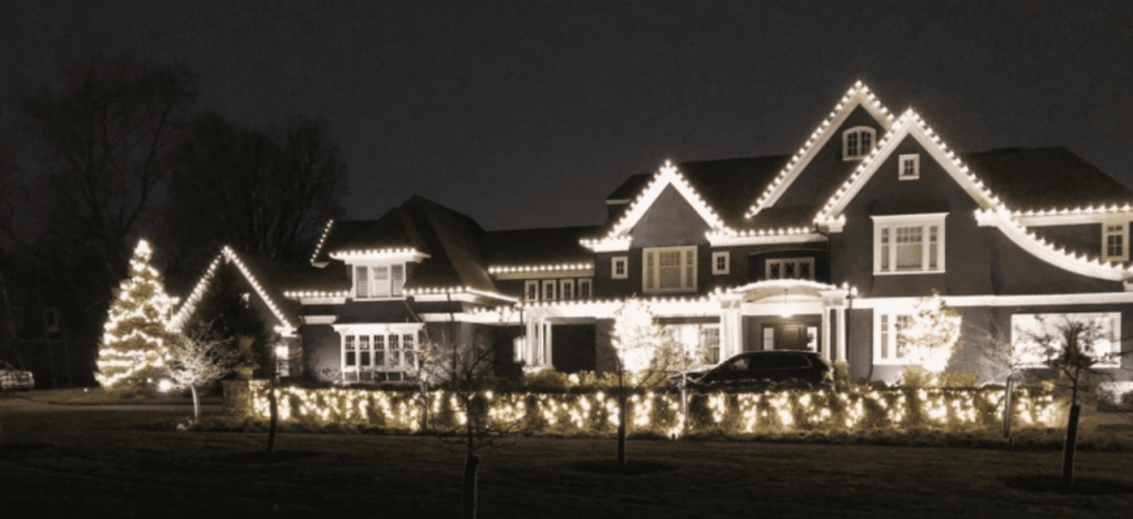 A grand house and its yard are beautifully illuminated with white holiday lights at night.