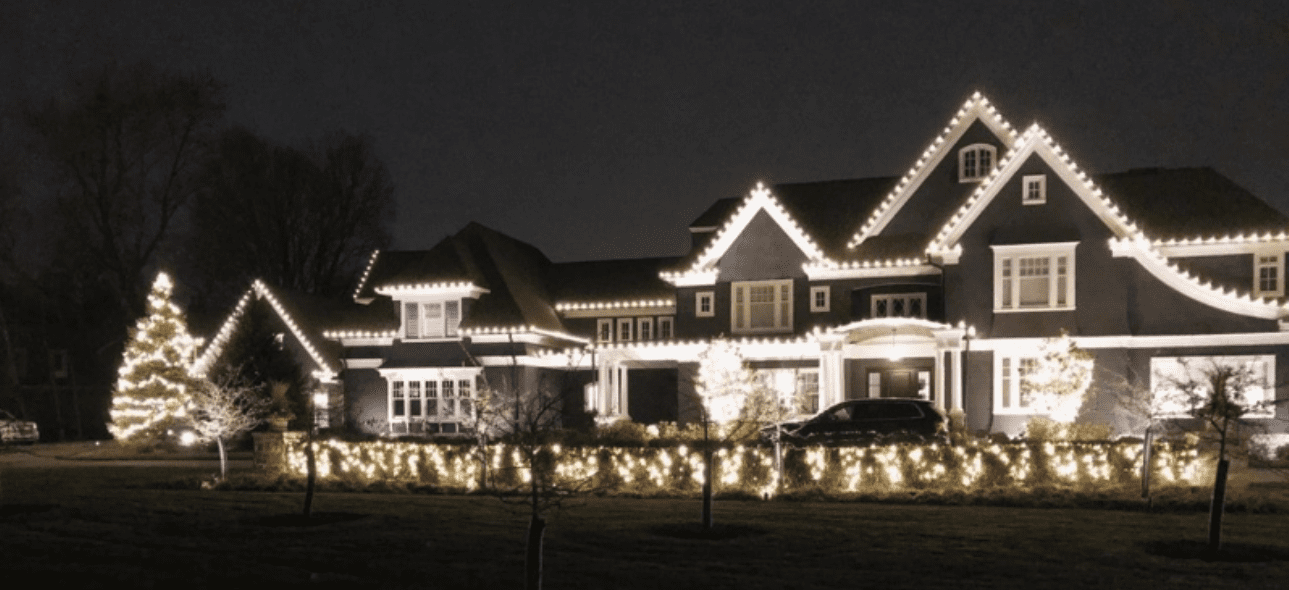 A grand house and its yard are beautifully illuminated with white holiday lights at night.