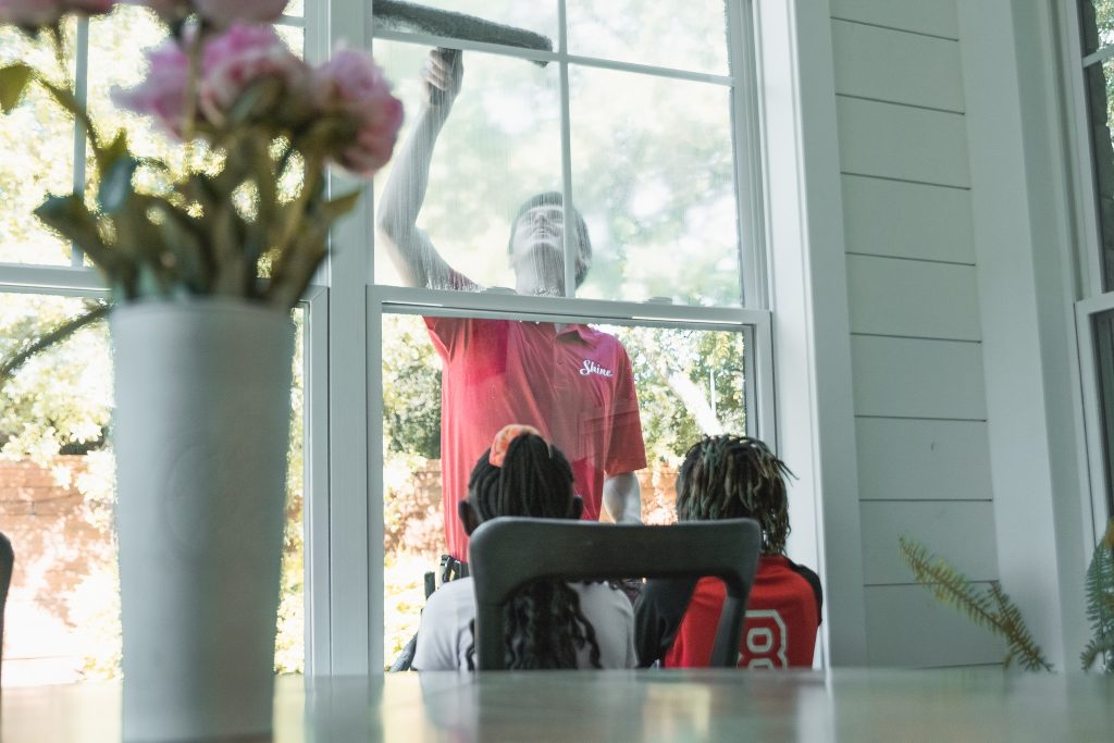 Man in red "Shine" shirt cleans a window, observed by two children