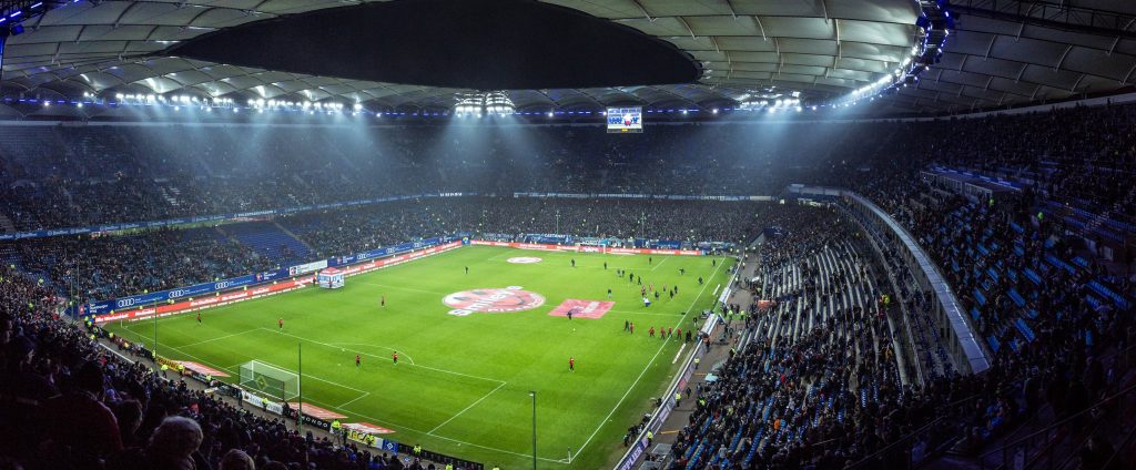 A vast, brightly lit football stadium filled with spectators and players on the green pitch
