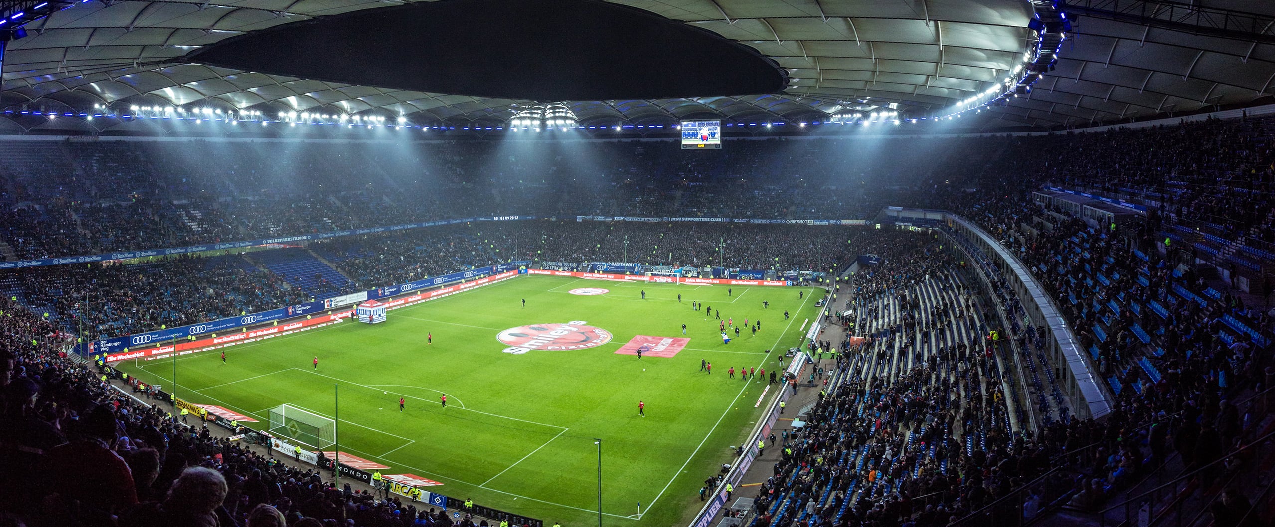 A vast, brightly lit football stadium filled with spectators and players on the green pitch