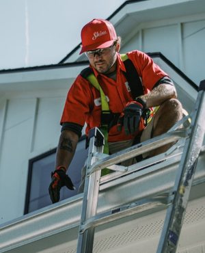 A worker in a red "Shine" hat and safety harness cleans a white house gutter from a ladder.