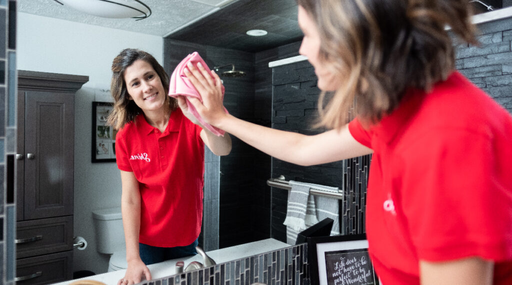A smiling woman in a red shirt cleans a bathroom mirror with a pink cloth, her reflection visible.
