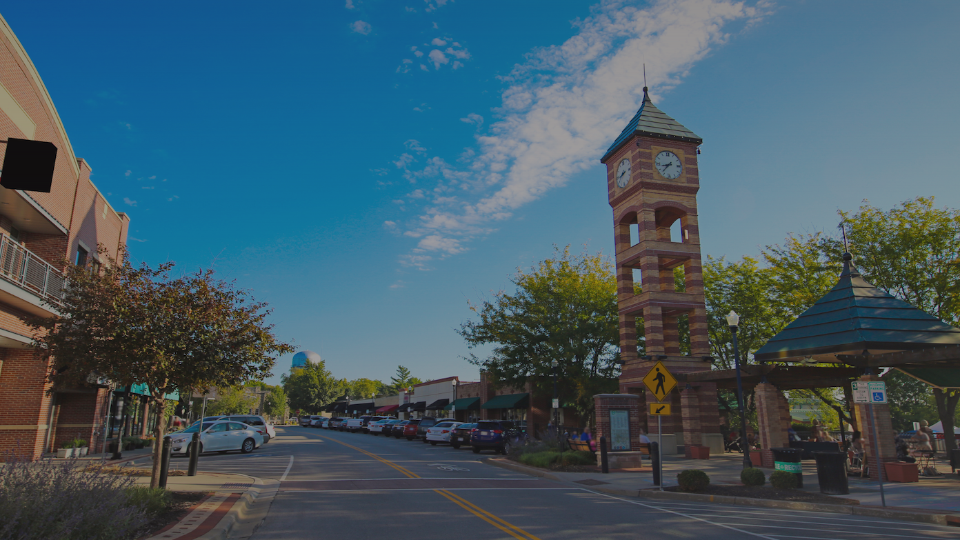 A sunny downtown street features a prominent brick clock tower, shops, and parked cars under a blue