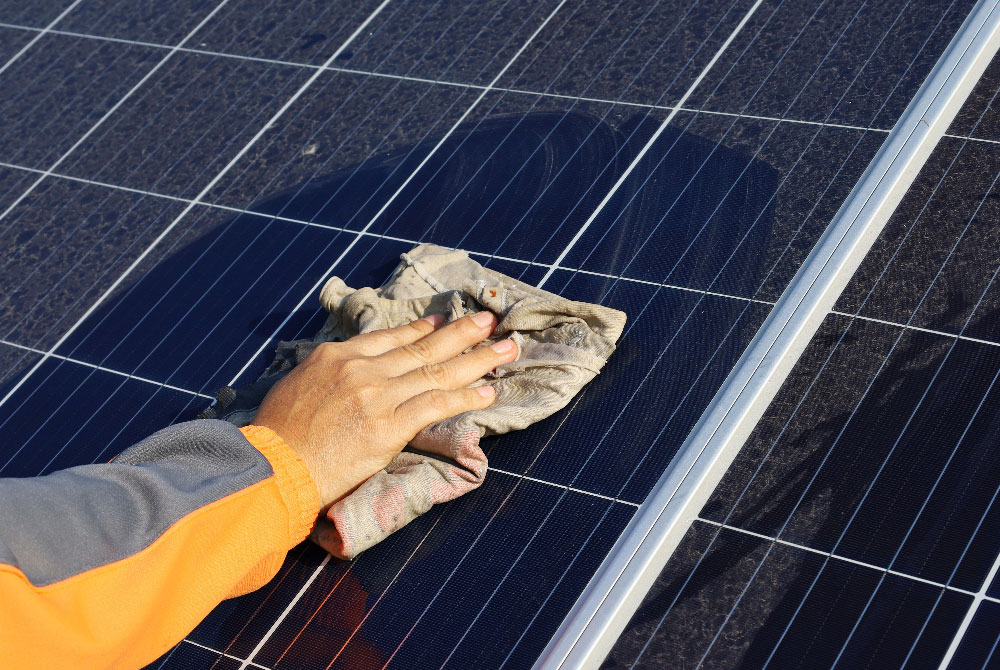 A hand in an orange sleeve cleans a dusty solar panel with a rag. TITLE: