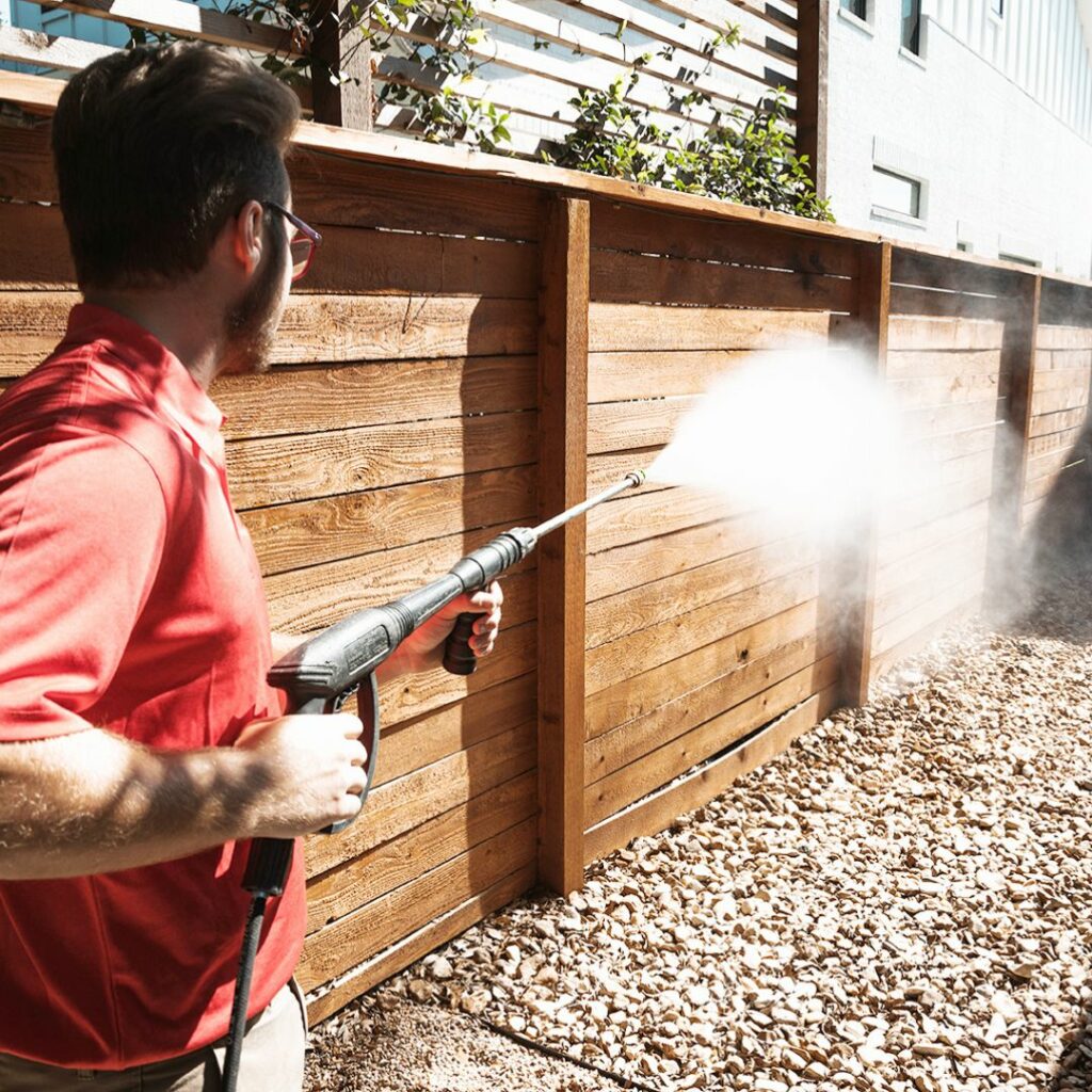 A man in a red shirt pressure washes a wooden fence, creating a spray of water.