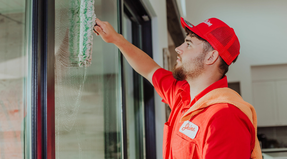 A bearded man in a red "Shine" uniform cleans a window with a green