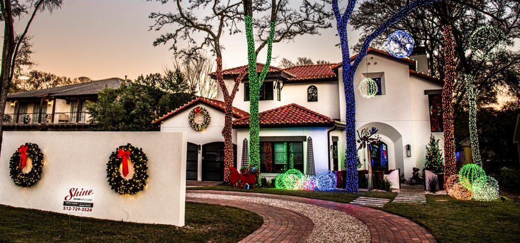 A large house is festively decorated with colorful Christmas lights on trees, wreaths, and