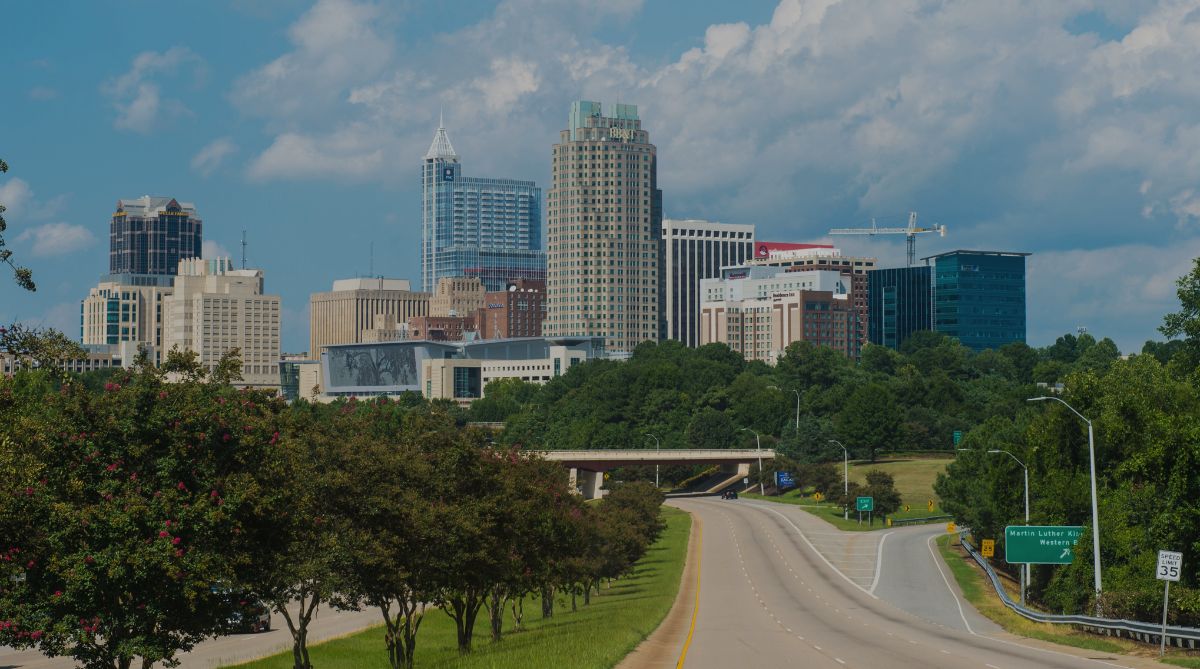 Raleigh's downtown skyline rises above a tree-lined highway under a blue sky with white