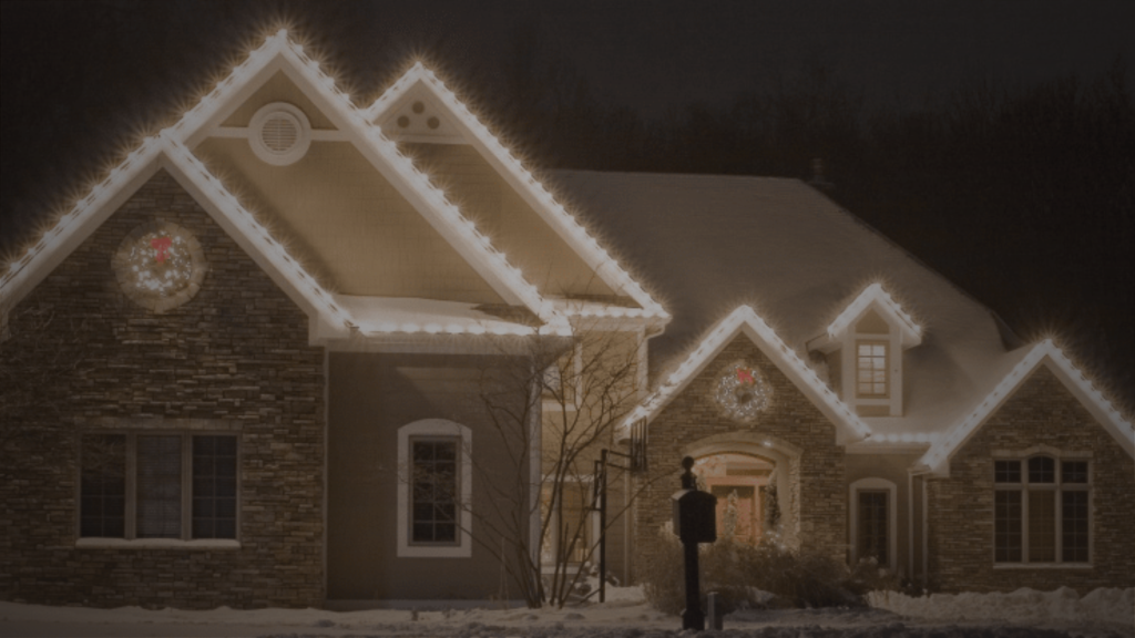 A snowy house glows with warm Christmas lights outlining its rooflines and gables at night.