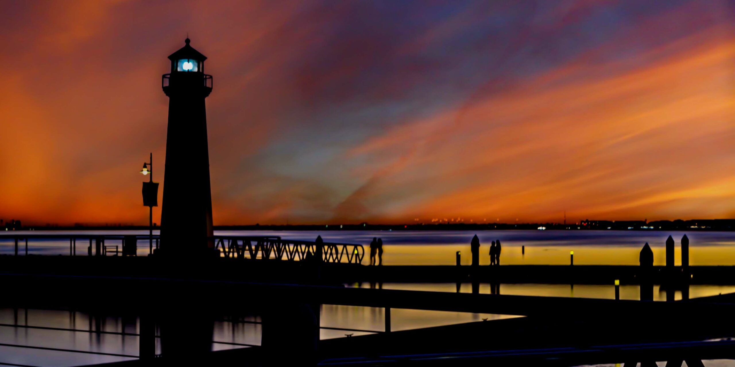 A lighthouse and pier are silhouetted against a vibrant, streaky orange and purple sunset