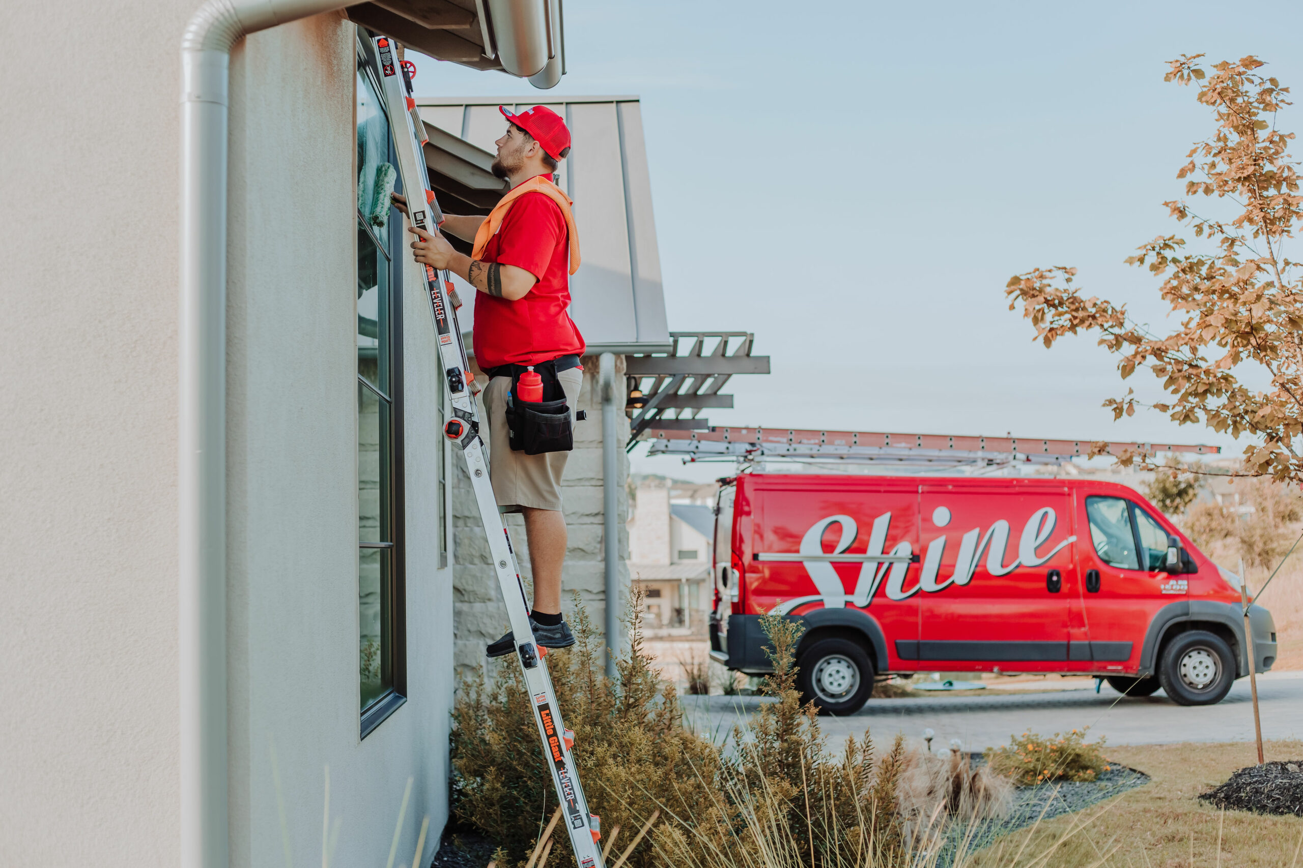 A man on a ladder cleans a house window while a red Shine service van waits nearby.