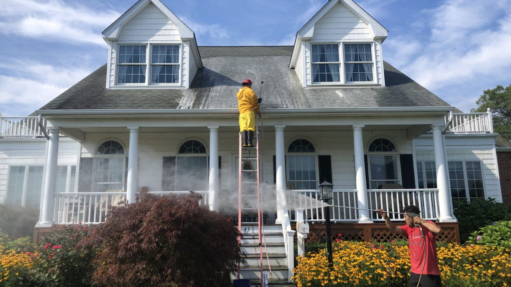 Workers in protective gear power wash a large white house, cleaning its dirty roof and porch.