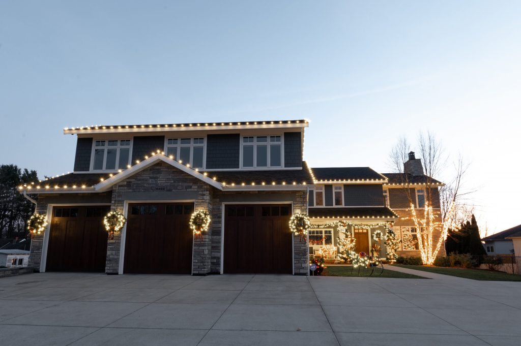 A large, modern house is brightly decorated with warm white Christmas lights outlining its roof and trees.