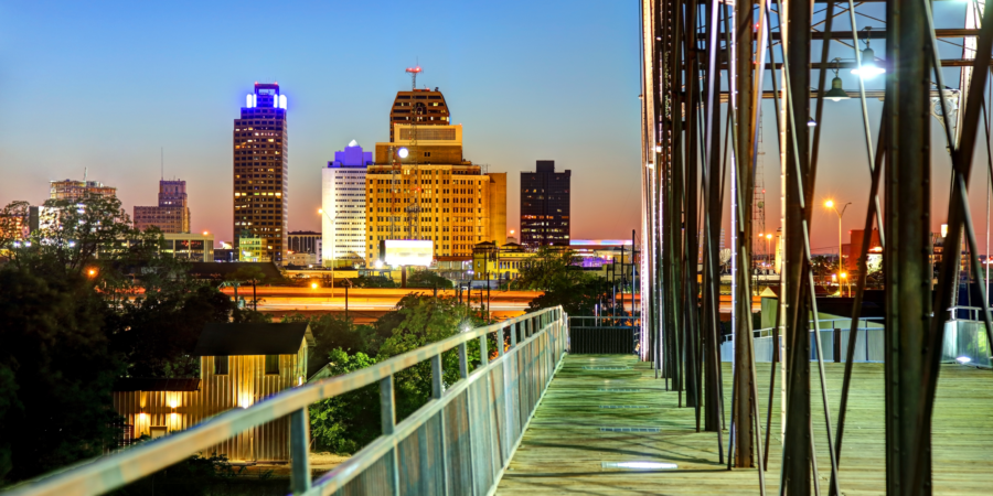 Dusk view of a vibrant city skyline with illuminated buildings and a pedestrian bridge in the foreground.