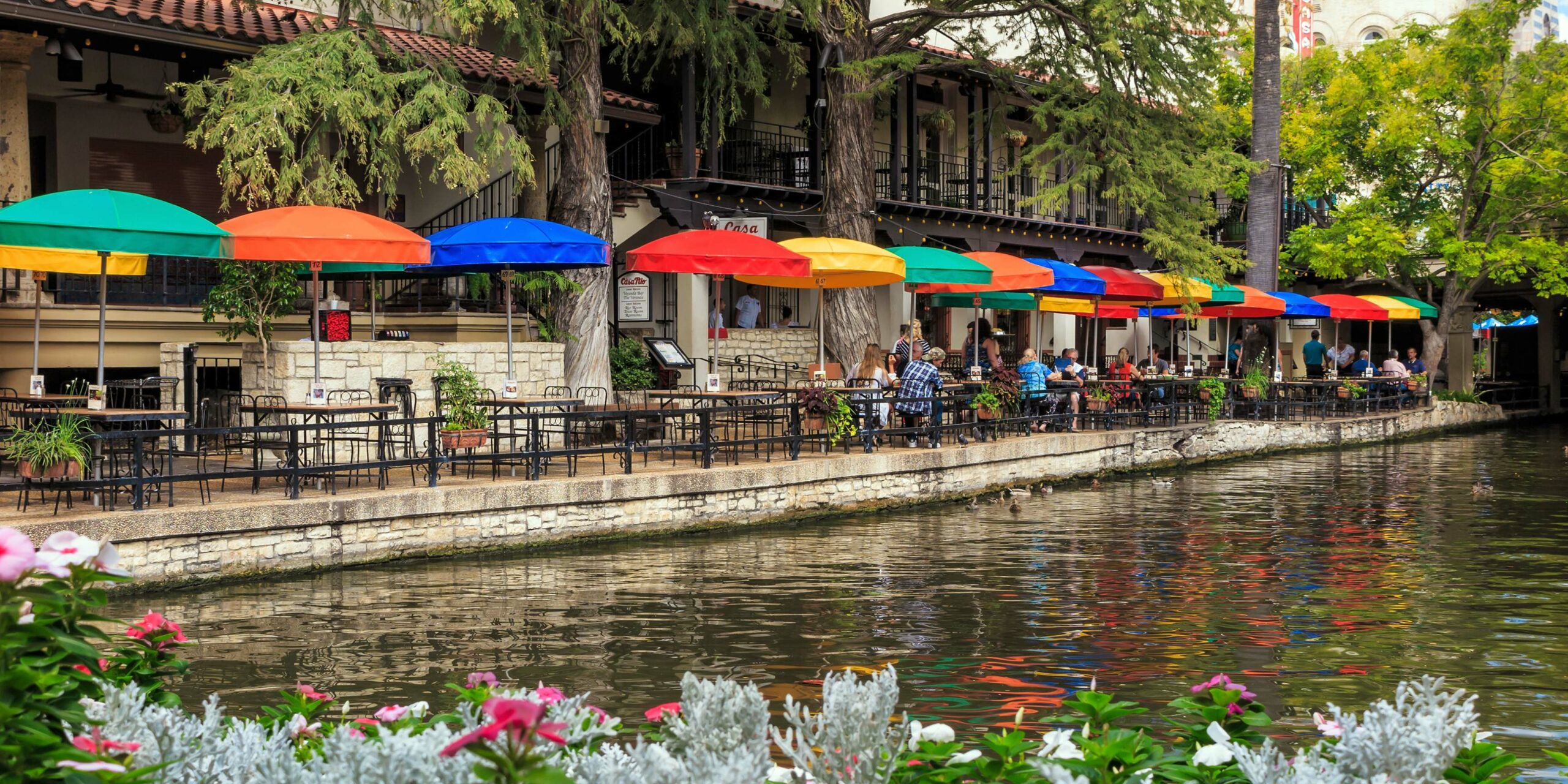 Colorful umbrellas shade diners enjoying a meal along a vibrant riverwalk lined with trees and flowers.