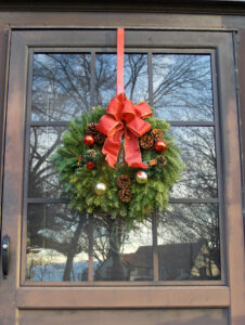 Holiday door wreath hanging from a red ribbon and hook over top a half-glass exterior front door.