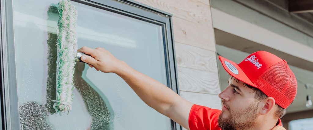 A person in a red 'Shine' cap cleans a window with a green washer.