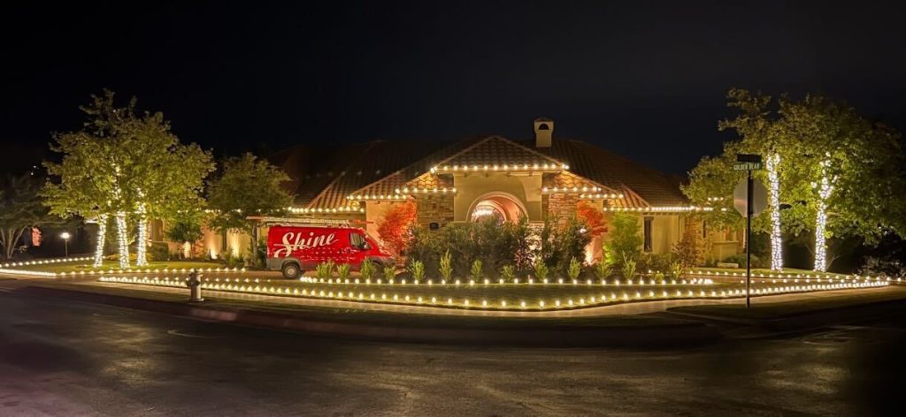 A large house and its trees are brightly decorated with warm white holiday lights at night, with a