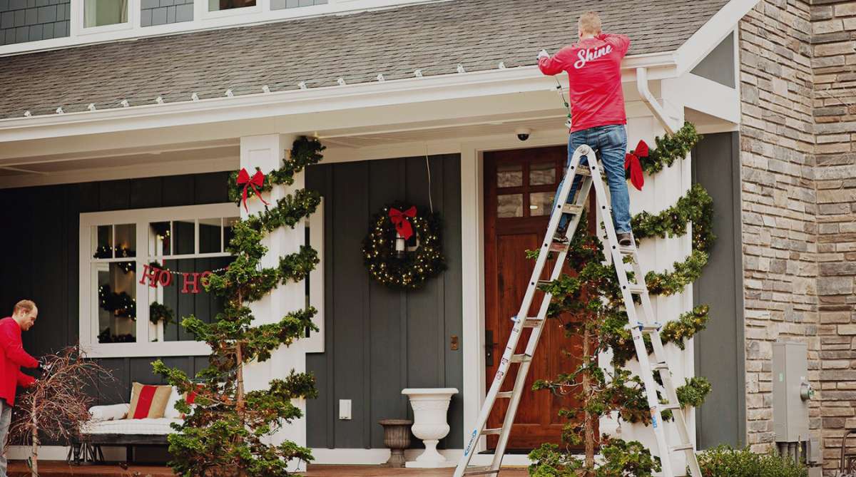 Two men decorate a house with Christmas lights and festive garland for the holidays.