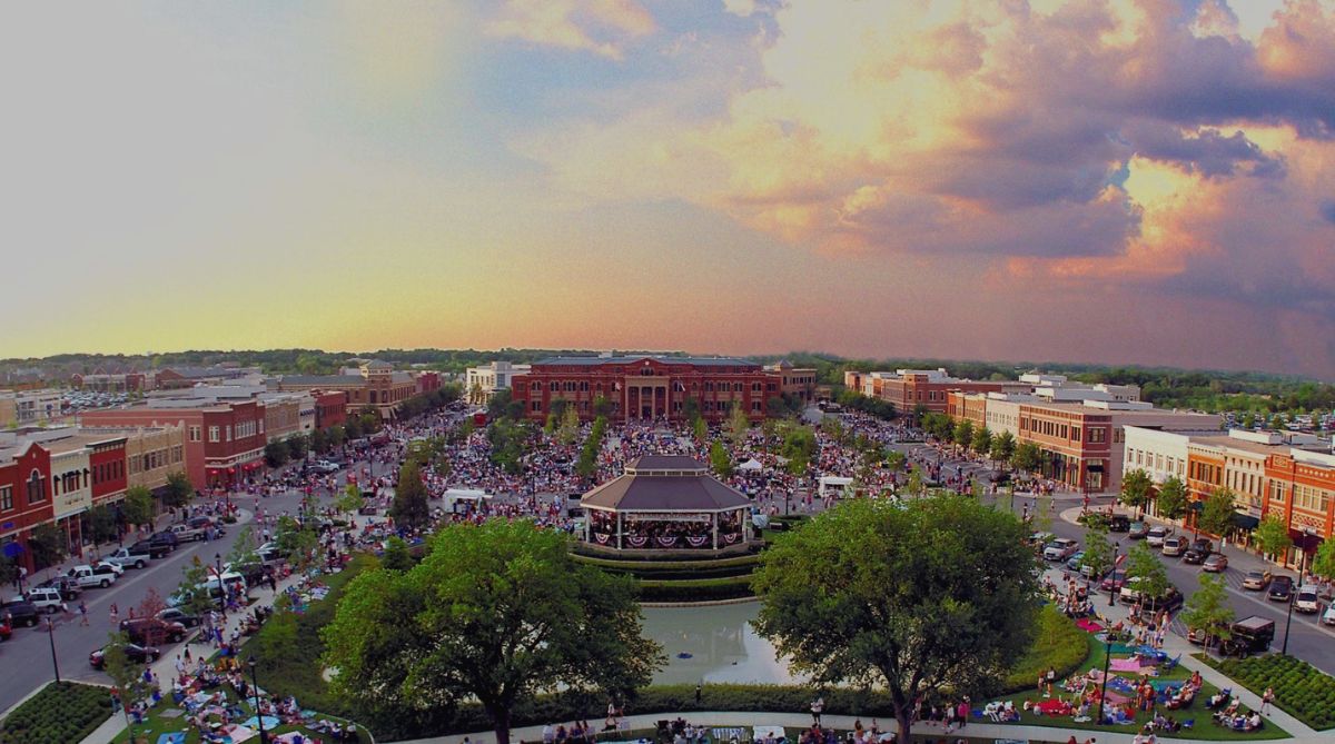 Crowds gather in a lively town square with a gazebo and pond under a colorful sunset.