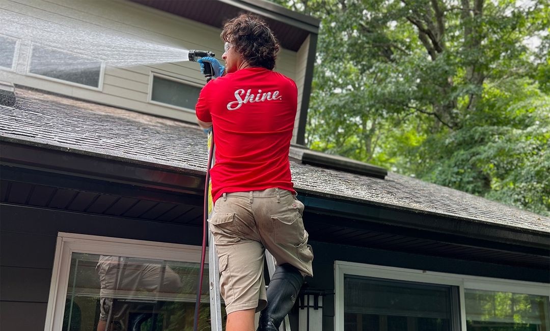 A worker wearing a red "Shine" shirt power washes the siding of a house from