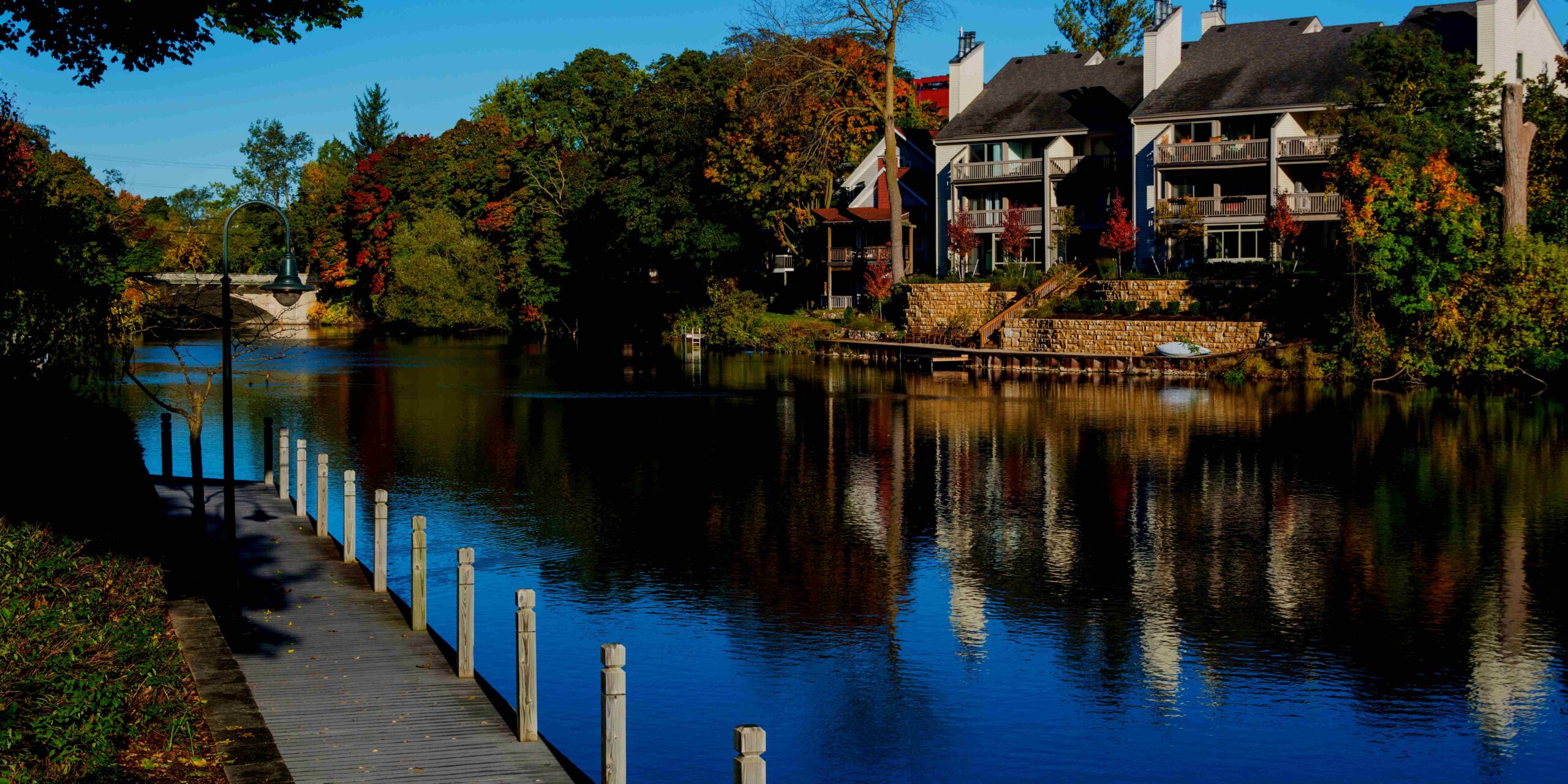 A wooden dock extends into a calm river reflecting waterfront buildings and colorful autumn foliage. TITLE: