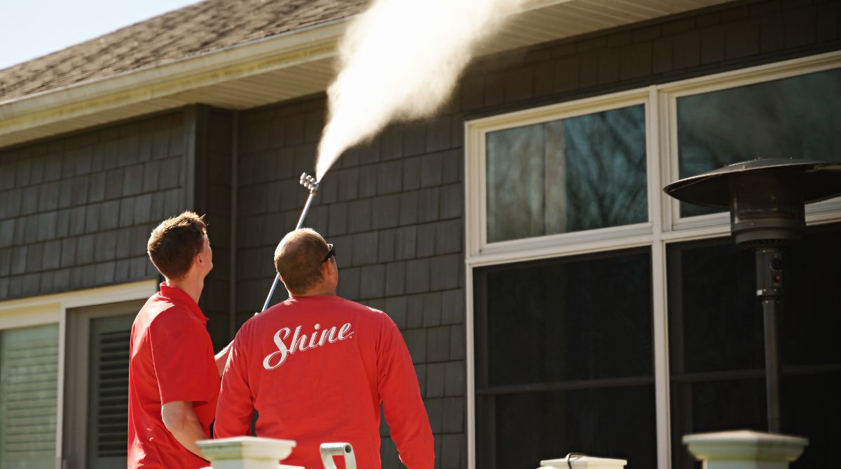 Two service workers in red shirts power wash the gutters and siding of a dark gray house.