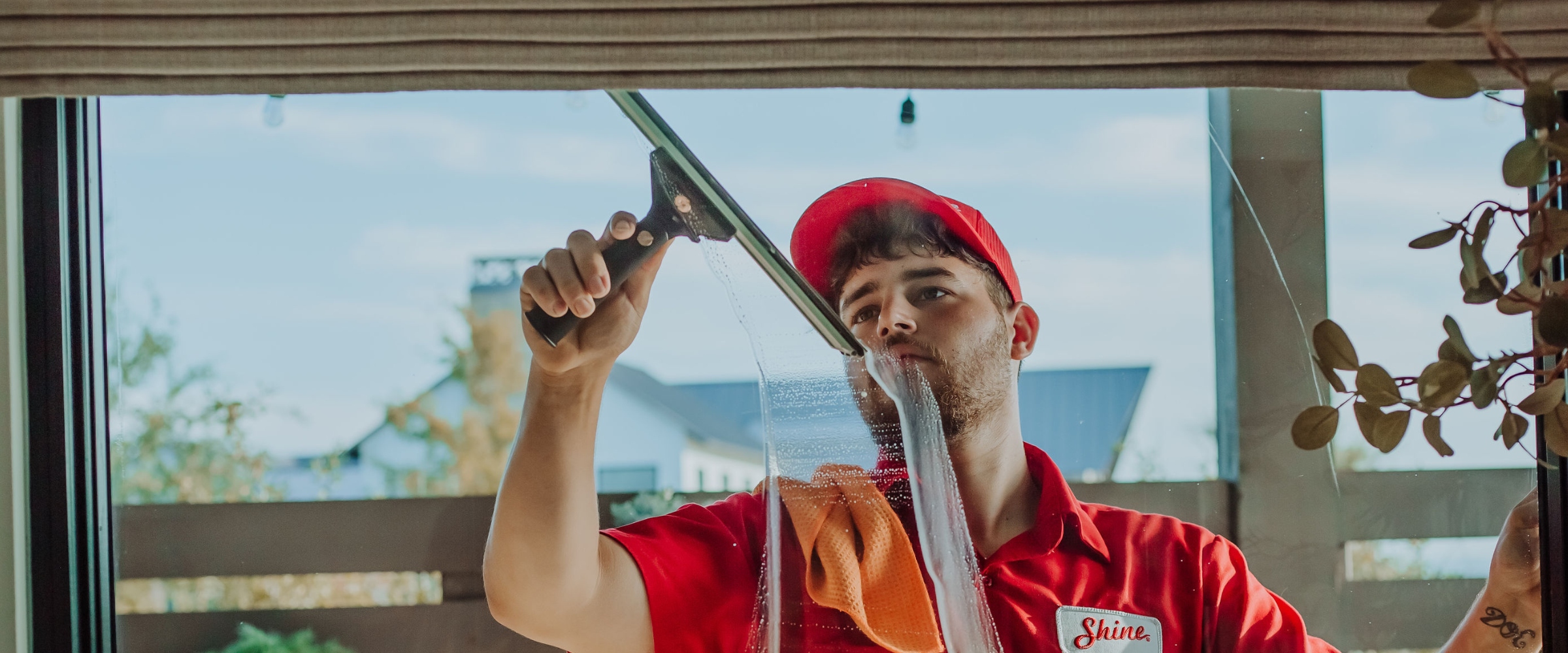 Man in red uniform with 'Shine' logo expertly cleans a large window with a sque