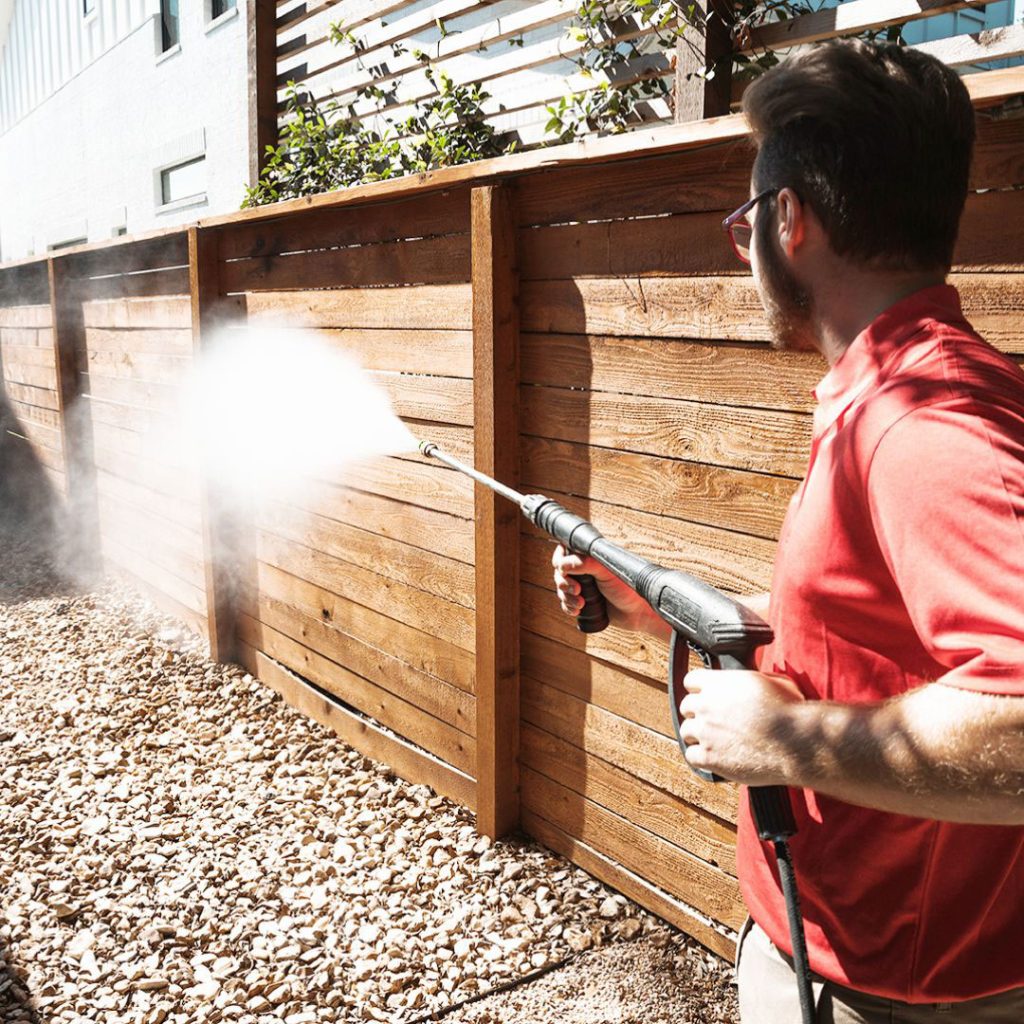 A man uses a pressure washer to clean a wooden fence, creating a misty spray. TITLE
