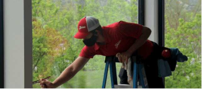 A masked window cleaner in a red uniform expertly cleans a large window pane from a ladder.