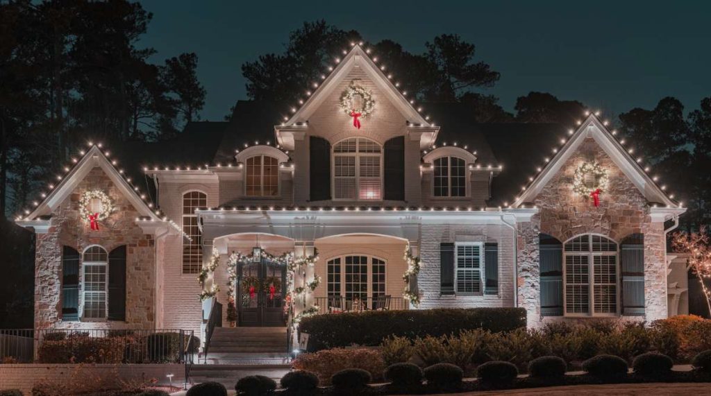 A large house is festively decorated with bright white Christmas lights and wreaths at night. TITLE