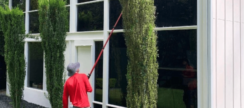 A person in a red shirt uses a long pole to clean tall windows on a white building