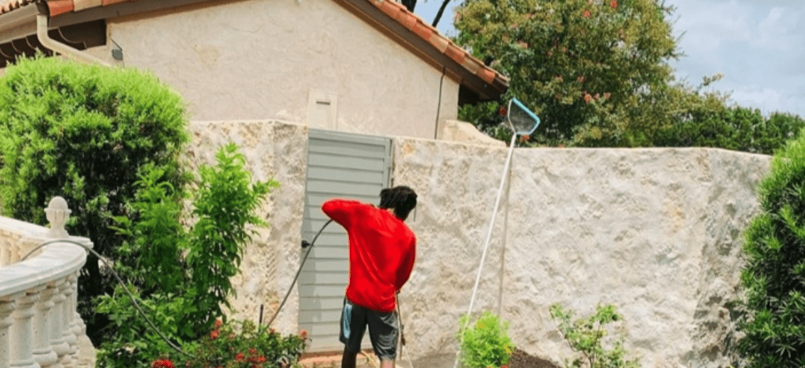A person in a red shirt pressure washes a textured stone wall next to green foliage. TITLE