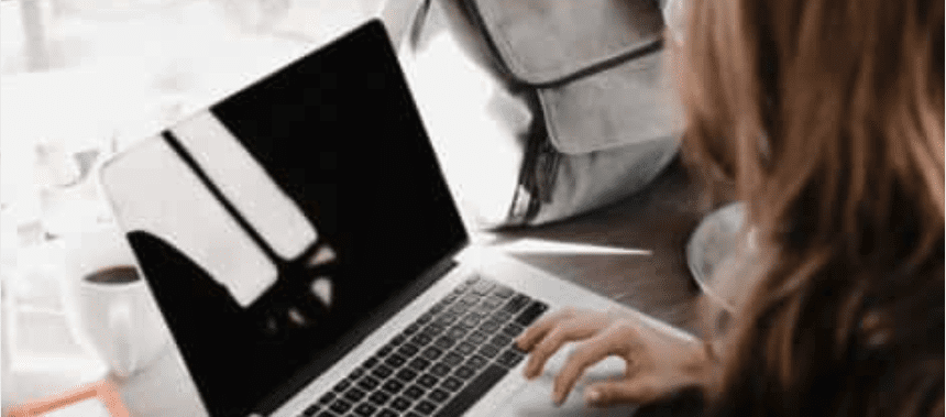 A person with brown hair works on a laptop, with a coffee cup and backpack nearby.