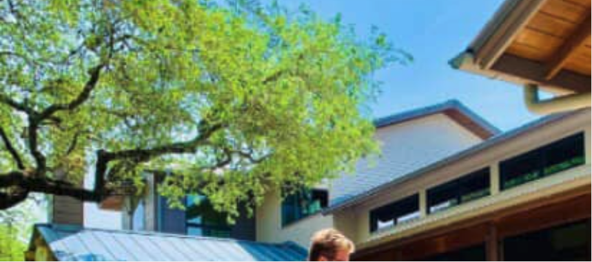 A large green tree shades a modern home featuring light walls and a dark roof under a blue sky