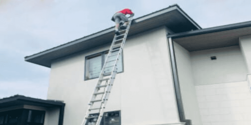 A person in a red hat climbs a tall ladder to reach the roof of a modern house.