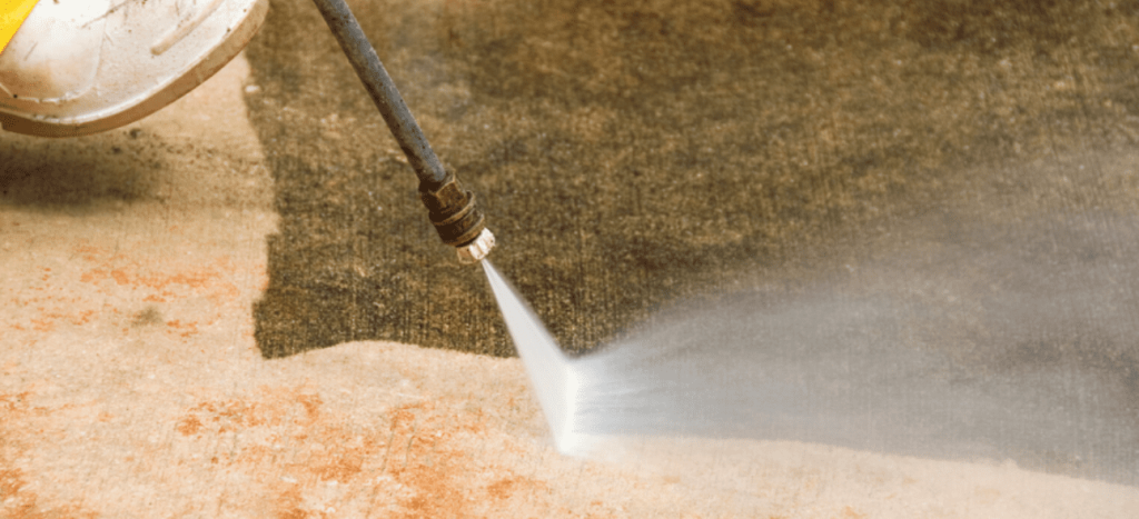 A person uses a pressure washer to clean a dirty outdoor surface, revealing a clean path.