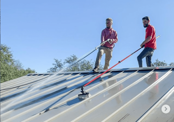 Workers power wash and brush a metal roof under a clear blue sky.
