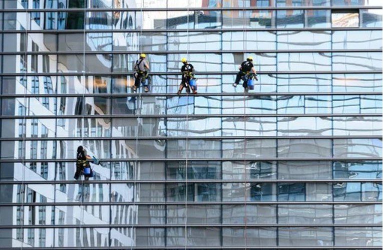 Four window washers rappel down a modern glass skyscraper, cleaning its reflective surface.