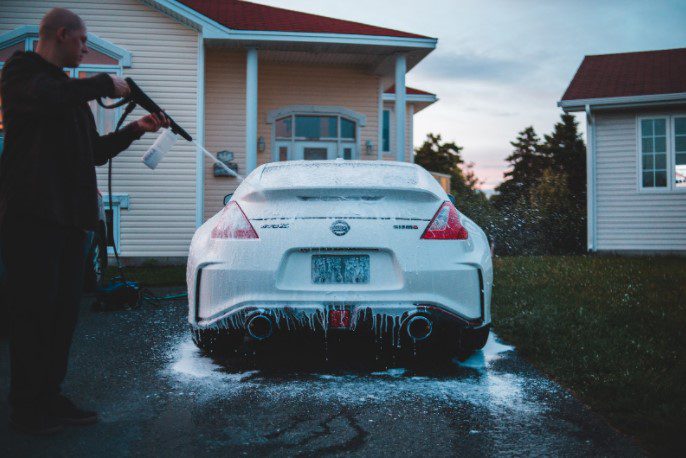 A man sprays a white Nissan sports car with soap foam from a pressure washer in a driveway