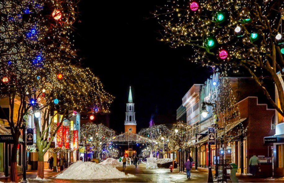 Downtown street at night adorned with sparkling holiday lights, colorful ornaments, and a prominent church steeple.