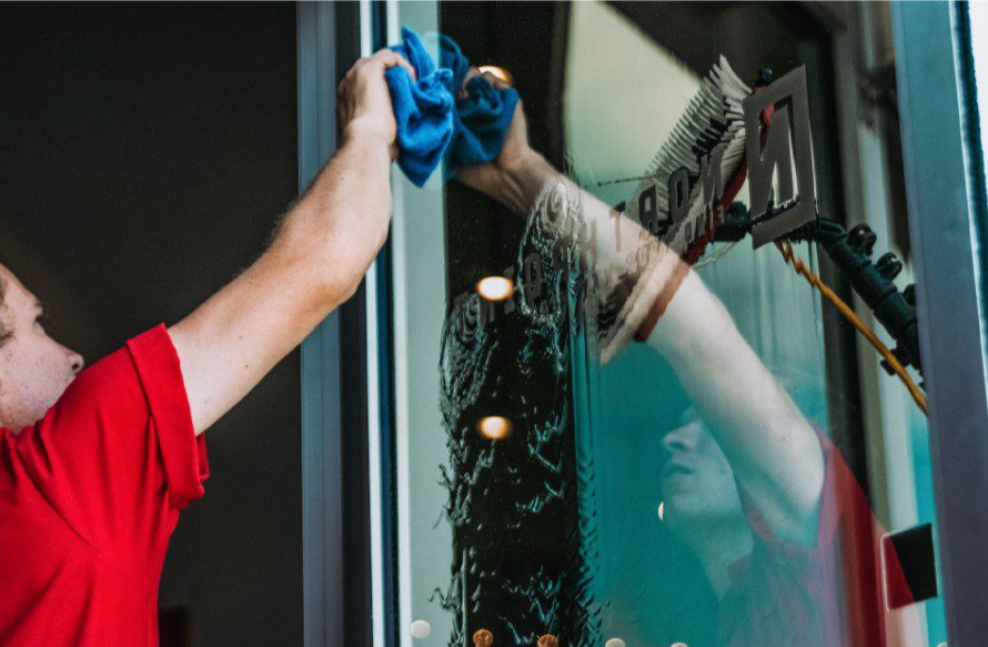 Man in a red shirt cleans a reflective window with a blue cloth. TITLE: